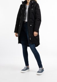 Sort parka-jakke med hætte, lynlås- og knaplukning, sidelommer og snoretræk. Båret sammen med blå jeans og grå sneakers.