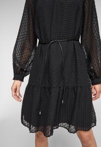 Black dress with textured fabric, sheer long sleeves, gathered tiers, elastic cuffs, and a thin black belt at the waist.