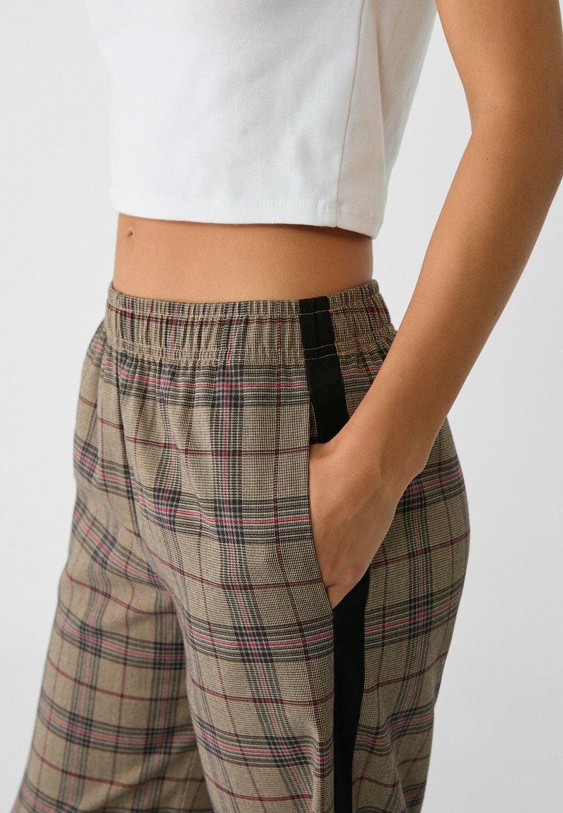 Pantaloni a quadri beige con linee rosse e nere, vita elasticizzata, tasche laterali e rifiniture nere lungo i lati. Top corto in cotone bianco.
