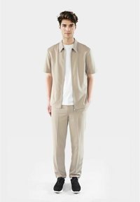 Beige kortermet skjorte med rett krage, parret med matchende bukser. Begge laget av glatt materiale, brukt med svarte sneakers.
