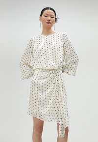 Vestido blanco con lunares negros, mangas largas, cintura ajustada y dobladillo asimétrico. Hecho de un tejido ligero y transparente con una textura suave.