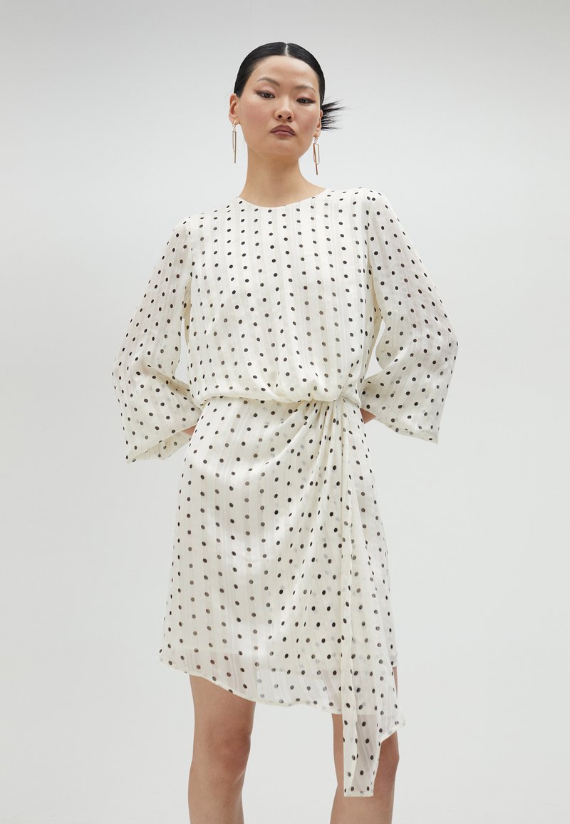 Vestido blanco con lunares negros, mangas largas, cintura ajustada y dobladillo asimétrico. Hecho de un tejido ligero y transparente con una textura suave.