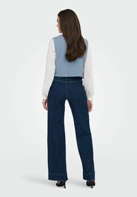Ljusblå kort topp med texturerat mönster, i kombination med mörk denim byxor med vida ben och fransad nederkant. Modell bär svarta högklackade skor.