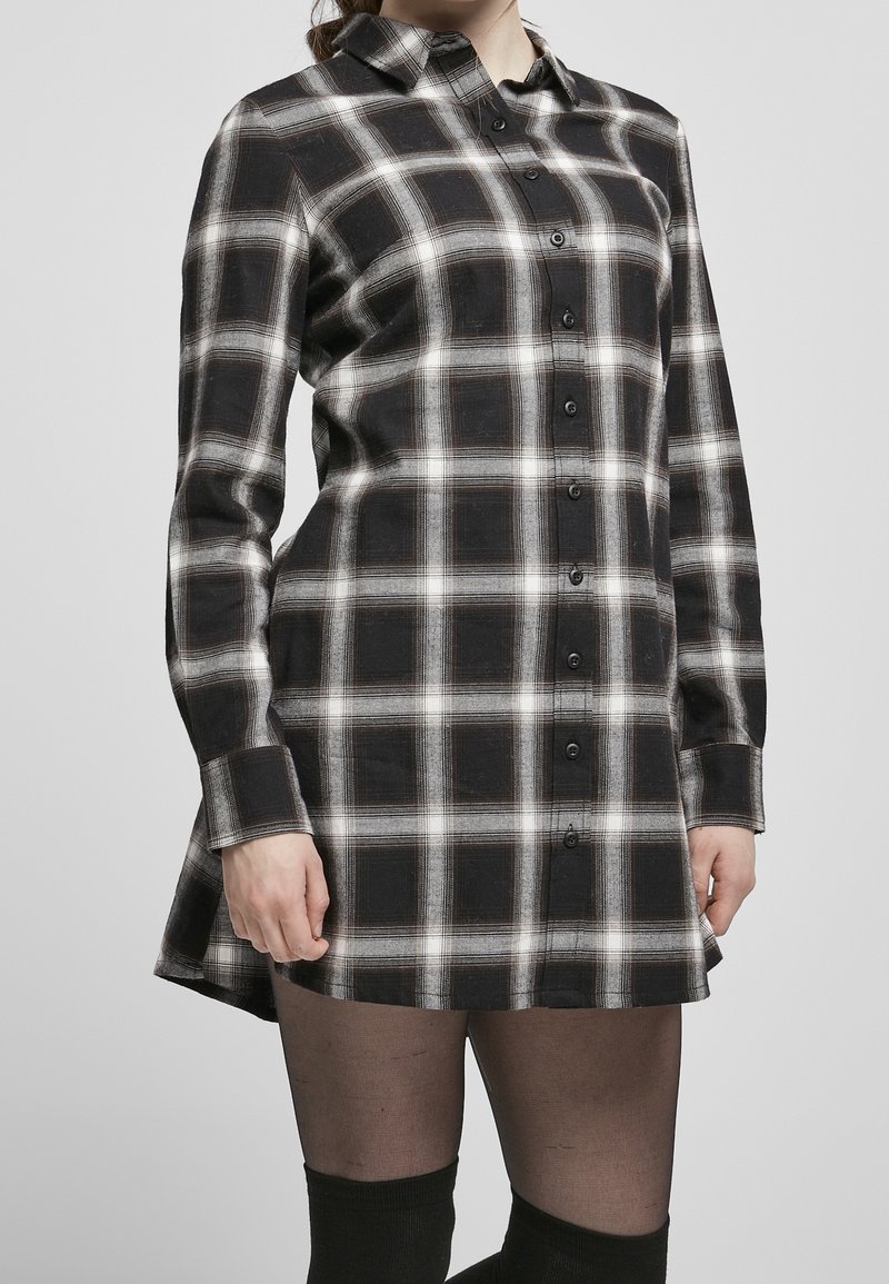 Femme portant une robe chemise à manches longues à carreaux noir et blanc, avec des collants noirs transparents et des chaussettes montantes.