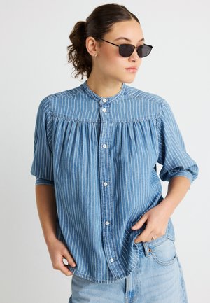 Blauwe denim blouse met witte verticale strepen, ronde kraag en knoopsluiting aan de voorkant, met een geplooid detail op de borst en driekwart mouwen.
