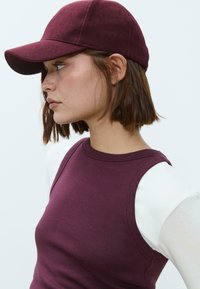 Burgundská manžestrová čiapka s výrazným krempou, vyrobená z textúrovanej tkaniny, so štruktúrovaným telom, nosená spolu s priliehavým, bezrukávovým topom.