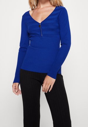 Frau trägt einen enganliegenden, langärmligen, blauen V-Ausschnitt-Pullover mit Reißverschlussdetail und schwarze Hose vor einfarbigem Hintergrund.
