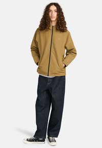 Junge Person mit langen lockigen Haaren, die eine beigefarbene Zip-Jacke, dunkle lockere Jeans und schwarze Turnschuhe trägt, steht mit den Händen in den Taschen.