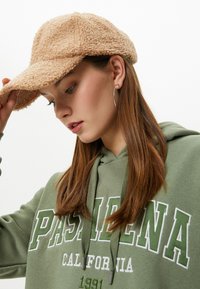 Gorra marrón esponjosa hecha de tela texturizada, combinada con una sudadera verde con letras blancas y cordones. Pendiente de aro plateado visible.