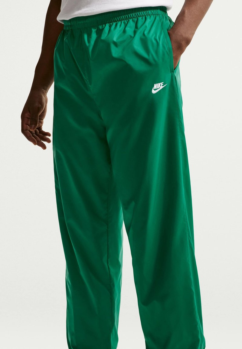 Mann trägt grüne Nike Trainingshose mit elastischem Bund und Hände in der Nähe der Taschen vor einfachem hellem Hintergrund.