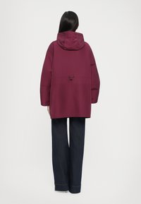 Veste d'extérieur bourgogne avec capuche et cordon de serrage. Design à coupe ample, associée à un pantalon en denim foncé avec des coutures visibles.