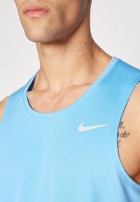 Svijetloplava sportska majica bez rukava s okruglim izrezom, sa bijelim Nike logotipom na lijevoj strani prsnog područja i glatkom, teksturiranom tkaninom.