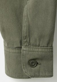 Gros plan sur un poignet en tissu vert olive avec des coutures visibles et un bouton vert foncé à quatre trous attaché près du bord.