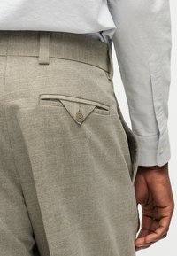 Κοντινό πλάνο στη μέση ενός ατόμου που φοράει μπεζ παντελόνια με ψαροκόκαλο και κουμπωμένη πίσω τσέπη, καθώς και ανοιχτό γκρι μακρυμάνικο πουκάμισο.