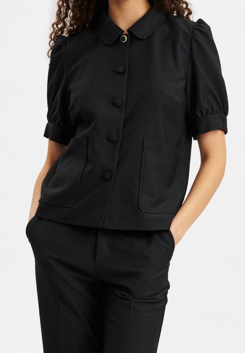 Schwarzes Kurzarmshirt mit rundem Kragen, Knöpfen vorne, zwei großen Taschen und einem glatten, strukturierten Stoff.