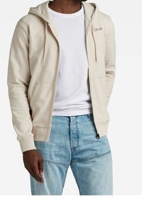 Felpa beige con zip, cappuccio regolabile con lacci, due tasche frontali e logo ricamato. Indossata sopra una t-shirt bianca e abbinata a jeans azzurri.