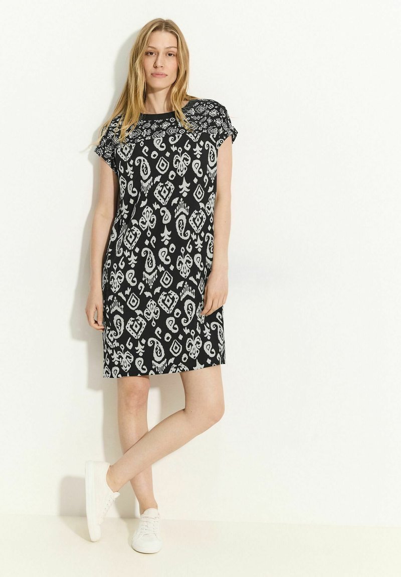 Schwarzes Kleid mit weißen Paisley- und geometrischen Mustern, kurzen Ärmeln und einem lockeren Schnitt, kombiniert mit weißen Sneakers.