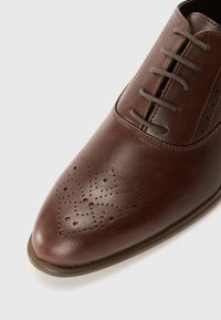 Brown leather dress shoe with a pointed toe, featuring decorative perforations and standard lacing. Smooth texture with a slight sheen.