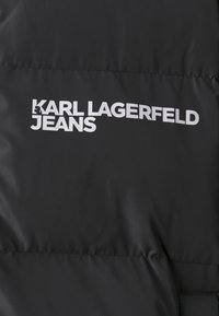 Svart polstret jakke med "KARL LAGERFELD JEANS"-logo i fet hvit tekst, som fremhever en glatt, shiny tekstur og minimalistisk design.