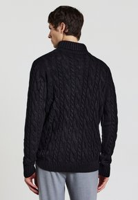 Maglione nero a maglia con un motivo a treccia testurizzato, un colletto alto e polsini e orlo a costine. Mostrato da dietro.