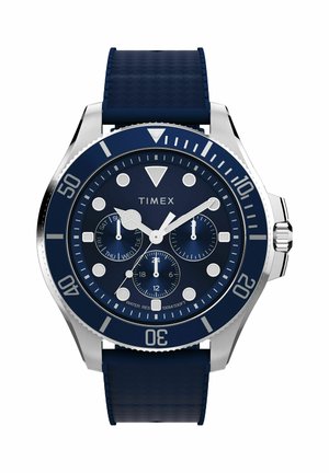 Kék Timex karóra ezüst kerettel, három segédtárcsával, amelyek a napot, a 24 órás időt és az órát mutatják, valamint texturált kék szilikon szíjjal.