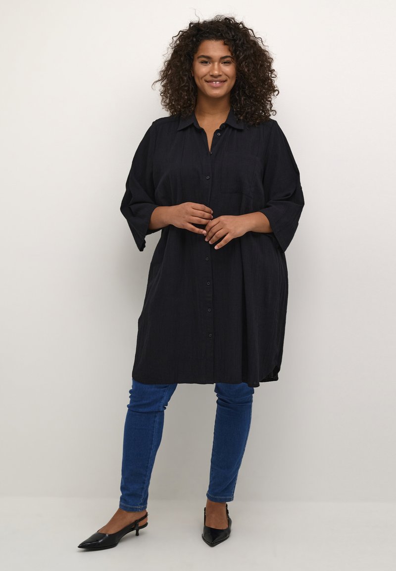 Robe chemise noire à boutons avec des manches trois-quarts, coupe décontractée, tissu texturé, poche poitrine unique, associée à un jean skinny bleu et des chaussures noires.