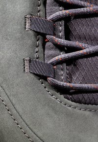Section de bottes en suédine grise avec des lacets gris foncé, des taches orange et des accents en tissu texturé entrelacés, avec des coutures détaillées visibles.
