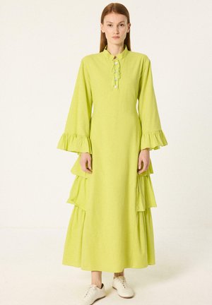 Femme debout portant une longue robe vert clair avec des manches évasées, des détails à volants sur les côtés et des chaussures blanches à lacets.