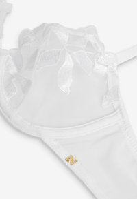 Balta mežģīņu bralette ar ziedu rakstu, ar caurspīdīgu augšējo audumu, gludu apakšējo daļu un mazu zelta emblēmu sānos.