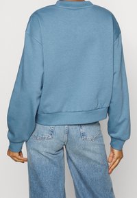 Sweat-shirt bleu clair à col rond et épaules tombantes. Poignets et ourlet côtelés. Porte avec un pantalon en denim bleu clair. Texture lisse.