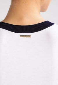 Bílá košile s námořnicky modrým žebrovaným kulatým výstřihem, na zádech malá zlatá logo destička, hladká textura látky, minimalistický design.