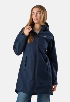 Junge Frau mit langen Haaren, die einen marineblauen Regenmantel mit Kapuze über Jeans trägt, den Kragen richtet und seitlich blickt, vor einem schlichten Hintergrund.