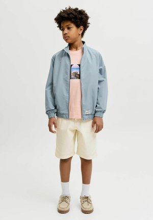 Junge steht und trägt eine hellblaue Zipjacke, ein pinkes Grafik-T-Shirt, cremefarbene Shorts, weiße Socken und beiges Schuhe vor einfachem Hintergrund.