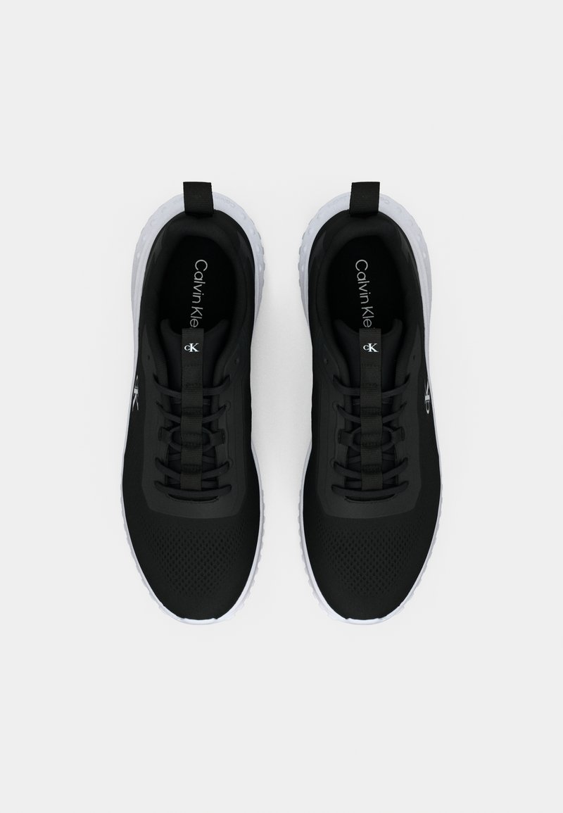 Une paire de baskets noires Calvin Klein avec des semelles blanches, une zone de orteils en mesh, des languettes de tirage et un design à lacets, vue de dessus sur un fond uni.