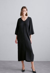 Vestido negro midi con escote en V, mangas anchas de tres cuartos y tela texturizada. Presenta un corte recto y pequeñas aberturas laterales.