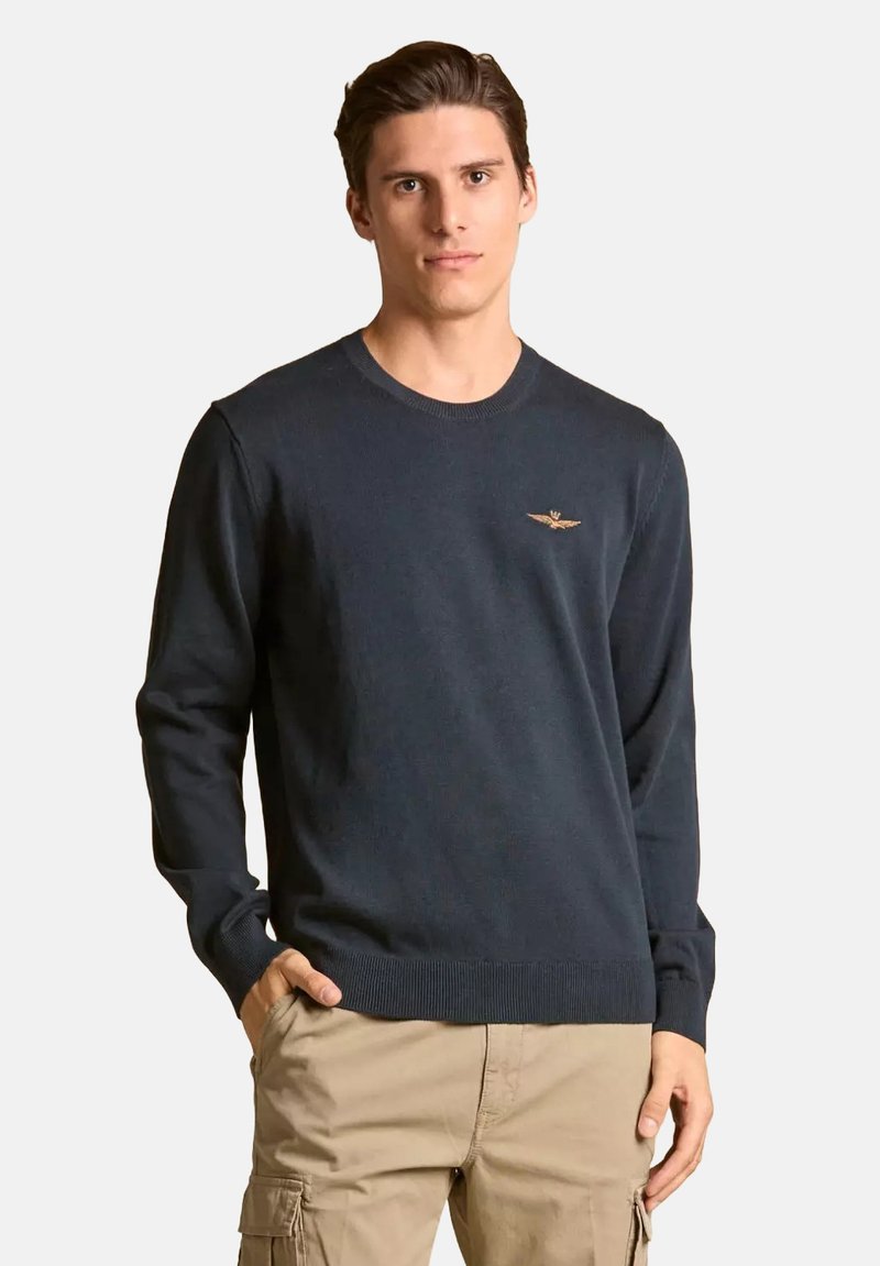 Maglione a collo rotondo di colore blu navy, realizzato con un tessuto liscio e dotato di polsini e orlo a costine. Presenta un piccolo logo ricamato in oro sul petto.