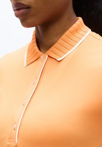 Gros plan d'une personne portant un polo pêche avec un col côtelé et une bordure blanche, mettant en valeur le col et la patte de boutonnage.