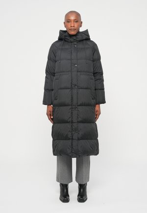 Doudoune noire avec capuche, longueur longue, design à panneaux, boutons-pression et poches avant, portée sur un pantalon gris et des bottes noires.