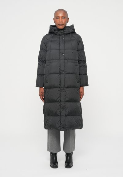 Giacca a vento nera con cappuccio, lunghezza lunga, design a pannelli, bottoni a pressione e tasche frontali, indossata sopra pantaloni grigi e stivali neri.