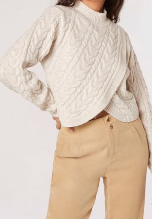 Kvinde iført en cremefarvet kabelstrikket sweater med asymmetrisk kant og beige bukser med høj talje, hånd på hoften, mod en ensfarvet baggrund.