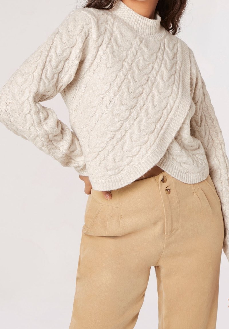 Femme portant un pull crème en maille torsadée avec un ourlet asymétrique et un pantalon beige taille haute, main sur la hanche, sur un fond uni.
