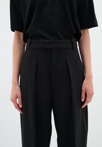 Black pleated trousers with a smooth texture, high waist, and belt loops. Paired with a loose-fitting black short-sleeve shirt.