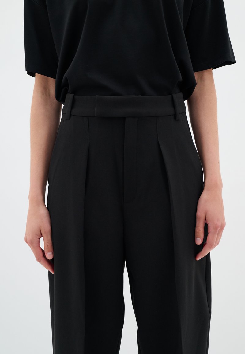 Schwarze, plissierte Hose mit glatter Textur, hoher Taille und Gürtel-Schlaufen. Kombiniert mit einem locker sitzenden, schwarzen Kurzarmhemd.