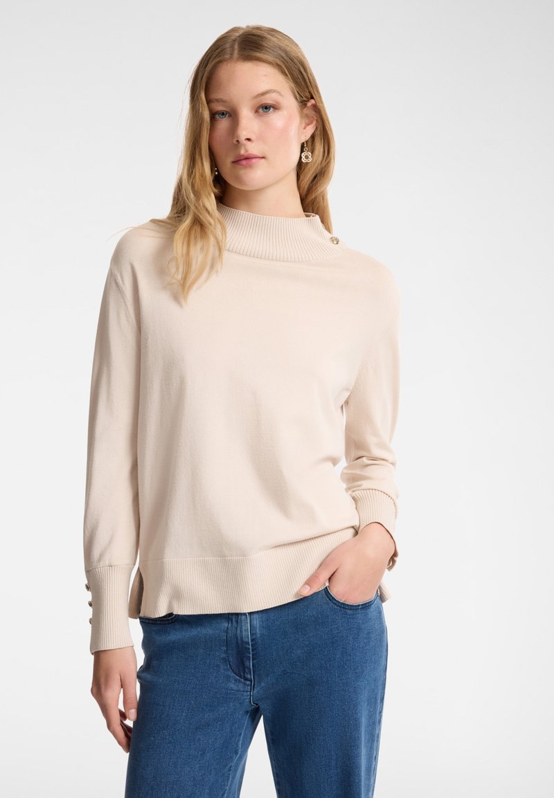 Vaalea beige poolikaulusneule, jossa on ribattu hihansuu ja helma, koristepainikkeet olkapäillä, yhdistettynä sinisiin farkkuihin.