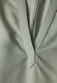 Tessuto verde pallido con design a V, dotato di cucitura centrale e dettaglio triangolare sul collo, texture morbida con una leggera lucentezza.