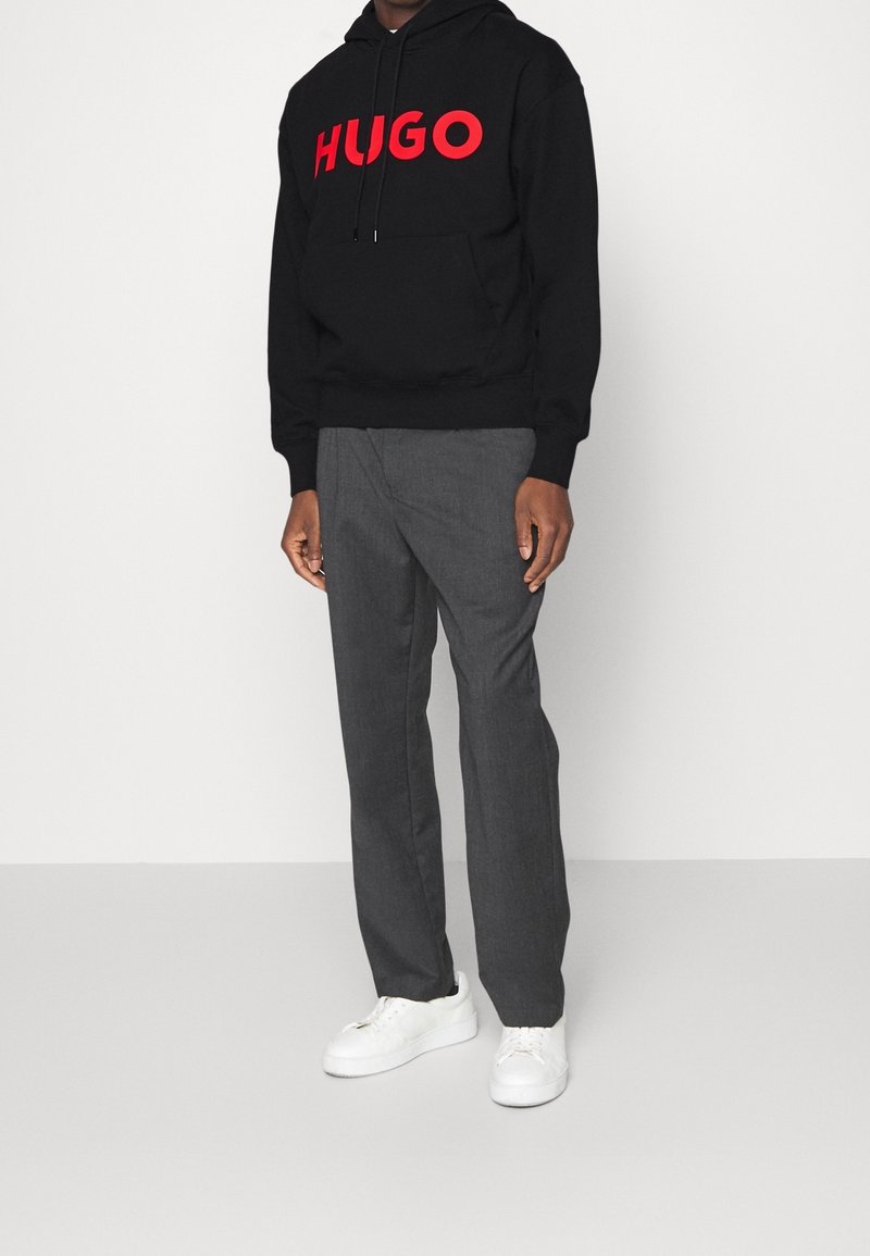 Svart hoodie med röd "HUGO"-logotyp, framfickan i kängurustil, parat med grå byxor och vita sneakers. Enkel, modern design.