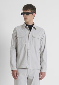 Camisa de botones de color gris claro con dos bolsillos en el pecho, textura acanalada y un corte relajado. Lleva puesta una camiseta negra debajo y se complementa con gafas de sol negras.