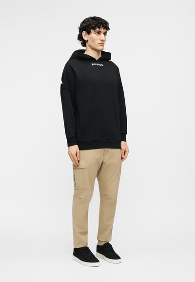 Noor mees seisab, kandes musta kapuutsiga pusa valge tekstiga, beežide taskupükste ja mustade tossudega, millel on valged tallad.