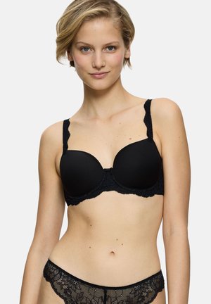 Donna che indossa un reggiseno in pizzo nero e mutandine in pizzo abbinate, rivolta in avanti con un leggero sorriso su uno sfondo chiaro e uniforme.