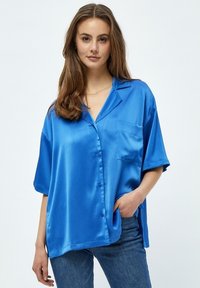 Camisa de satén azul de manga corta, con cierre de botones y bolsillo. Textura suave y corte relajado. Combinada con jeans azules.